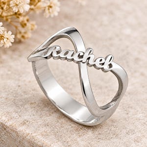 Peut inclure: Une bague en métal argenté poli présentant un motif en forme de symbole infini. Le prénom Rachel est inscrit en écriture cursive au centre de la boucle infinie, intégré directement dans la structure de l'anneau de la bague.