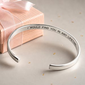 Puede incluir: Una pulsera de brazalete de metal plateado con un diseño abierto y un acabado pulido. La superficie interior de la pulsera está grabada con el texto en negro, I WOULD FIND YOU IN ANY LIFETIME, en una fuente clara y sin remates.