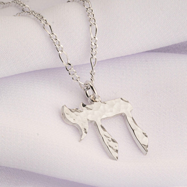 Pu&ograve; includere: Una collana color argento con un ciondolo a forma di lettera ebraica Chai. Il ciondolo presenta una superficie martellata e testurizzata ed &egrave; sospeso a una delicata catena color argento con maglie ovali allungate dal riflesso lucido.