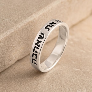 May include: A silver ring with Hebrew text engraved around the band. The ring is set against a neutral background. The text is in a dark color, contrasting with the shiny silver metal.