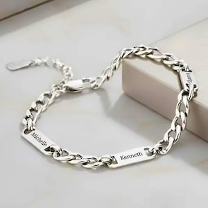 Puede incluir: Una pulsera de eslabones plateados con tres placas metálicas rectangulares. Cada placa está grabada con un nombre, incluyendo Michelle, Kenneth y un tercer nombre parcialmente visible. La pulsera incluye un cierre de mosquetón y una cadena extensora.