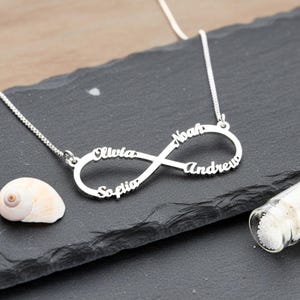 May include: A silver-toned infinity symbol necklace featuring the names Olivia, Noah, Andrew, and Sofia integrated into the metal loops. The pendant is attached to a delicate silver chain, creating a continuous design with cursive lettering.