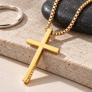 May include: A gold-coloured cross pendant necklace featuring a box chain and a smooth, polished finish. The vertical bar of the cross is engraved with the name DAVID in a clean, sans-serif font. A simple, plain silver-coloured ring sits nearby.