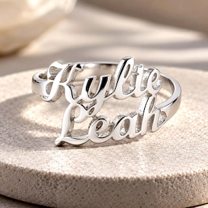 May include: A polished silver-toned ring featuring two stacked, cursive-style names, Kylie and Leah, integrated into the band design. The metal has a reflective, smooth finish, and the lettering is clearly legible in a script font across the front.
