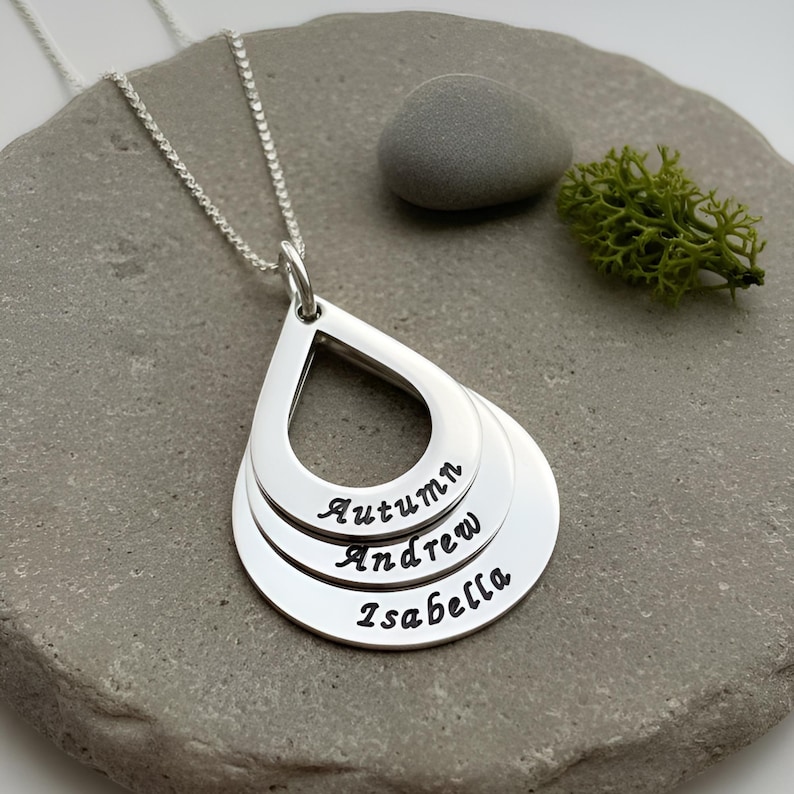 May include: A silver necklace featuring three stacked, teardrop-shaped pendants hanging from a delicate chain. Each pendant is engraved with a name in black script: Autumn, Andrew, and Isabella, arranged from the smallest top layer to the largest base.