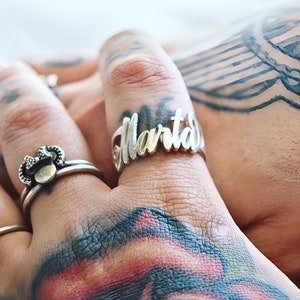 May include: A silver ring with the name "Marta" engraved on it. The ring is worn on the index finger of a hand with tattoos.