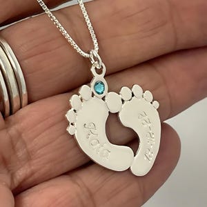 Puede incluir: Collar de plata con un dije de dos huellas de pies de bebé entrelazadas. Las huellas están grabadas con los nombres "Ava" y "Ezra" y una pequeña piedra preciosa azul está engastada en el centro.