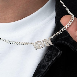 Puede incluir: Collar plateado con el nombre "KEVIN" en letras mayúsculas. La cadena es de estilo curb. El collar se muestra sobre una camisa blanca y una chaqueta negra.