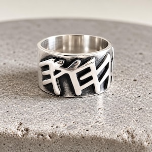 May include: A wide silver band ring featuring a polished finish with recessed, darkened areas that highlight a series of angular, runic-style characters embossed around the exterior surface of the metal band.