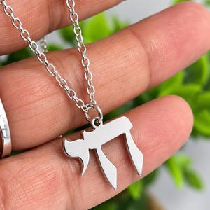 Könnte beinhalten: Eine silberfarbene Metallhalskette mit einem polierten Anhänger in Form des hebräischen Chai-Symbols. Der Anhänger hängt an einer zarten, passenden silberfarbenen Gliederkette und zeichnet sich durch eine glatte, reflektierende Oberfläche aus.