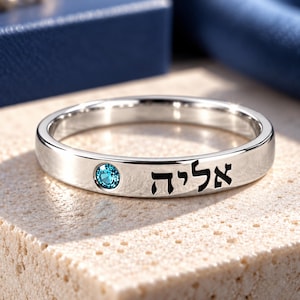 May include: A polished silver-toned band ring featuring a single round, bright blue gemstone set into the metal surface next to black Hebrew lettering that reads אליה. The ring has a smooth, reflective finish and a simple, clean design.
