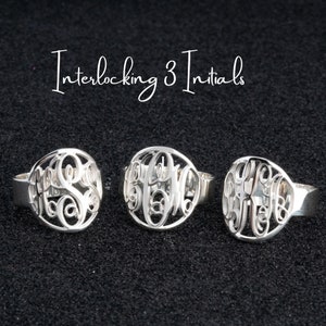 Könnte beinhalten: Drei silberne Ringe mit ineinander verschlungenen Initialen. Die Ringe liegen auf einem schwarzen Hintergrund. Interlocking 3 Initials.