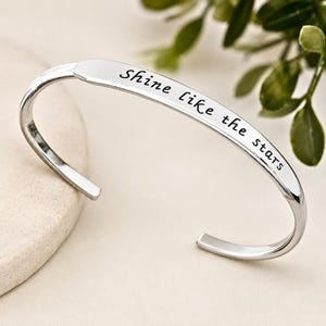 Puede incluir: Una pulsera tipo brazalete de metal plateado con acabado pulido y diseño abierto. La superficie frontal de la pulsera está grabada con el texto negro en cursiva que dice, Shine like the stars, centrado a lo largo de la banda curva.