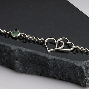 Op de afbeelding: Een zilveren kettingarmband met een groene edelsteen en een dubbele hartbedel.