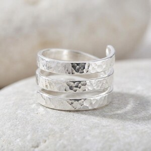 May include: A silver ring with a hammered finish. The ring has a spiral design, with three bands wrapping around the finger. The top band curves upwards, creating an open-ended look. The ring is displayed on a light-coloured surface.