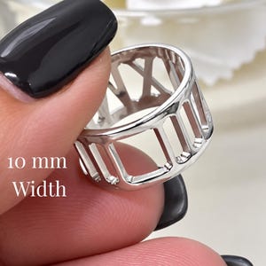 May include: A silver-toned ring featuring a cut-out design of Roman numerals around the band. The ring has a polished metallic finish and a width of 10 mm as indicated by the text printed on the image. The band is open-work with vertical bars.