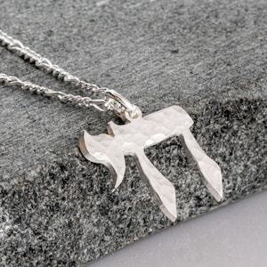 Pu&ograve; includere: Un ciondolo color argento a forma di carattere ebraico Chai, caratterizzato da una finitura martellata e testurizzata sulla superficie. Il ciondolo &egrave; fissato a una delicata collana a catena in metallo color argento, creando un aspetto metallico riflettente.