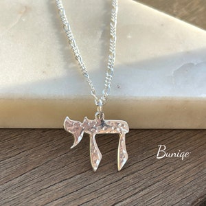 Pu&ograve; includere: Una collana in argento con un ciondolo a forma di simbolo ebraico Chai. Il ciondolo presenta una finitura martellata e testurizzata ed &egrave; appeso a una sottile catenina d'argento. La parola Buniqe &egrave; stampata in bianco nell'angolo in basso a destra.