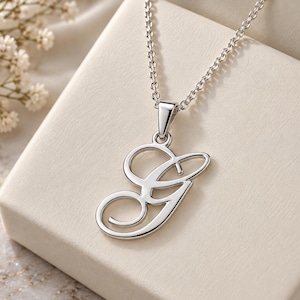 May include: A silver-toned necklace featuring a cursive letter G pendant. The pendant is suspended from a delicate silver-toned chain. The design consists of a polished metallic finish with smooth, flowing lines that form the uppercase letter G.