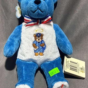 May include: A blue plush bear with a white and blue bow tie and a white shirt with a bear in a postal worker uniform. The bear has a tag attached. The bear's feet have a mailbox design.