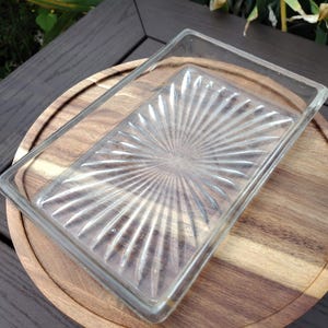 May include: A clear, rectangular glass dish with a sunburst pattern sits atop a round wooden serving tray. The tray is placed on a dark brown wooden surface, creating a contrast of textures and colors. The dish is ideal for serving appetizers or desserts.