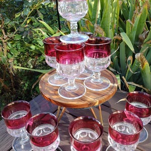 May include: A collection of vintage wine glasses with ruby red rims and clear, patterned bowls. The glasses are arranged on a small wooden table and a wooden surface, with greenery in the background. The stemware is likely from the mid-century.