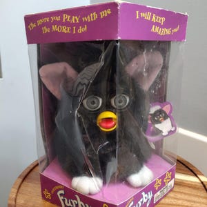 May include: A black Furby toy in its original box. The Furby has large, expressive eyes, a yellow beak, and pink ears. The box is purple and features the Furby logo and text that reads "The more you PLAY with me the MORE I do!" and "I will KEEP AMAZING you!"