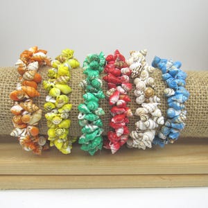 May include: Six bracelets made of small, spiraled seashells in vibrant colors including orange, yellow, green, red, white, and blue. Each bracelet is constructed from multiple tiny shells clustered together to form a textured, circular band.