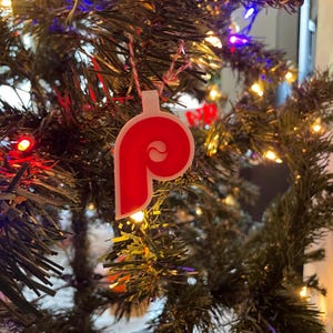 May include: A red and white Christmas ornament shaped like the letter "P" with a small circle inside the letter. The ornament is hanging on a green Christmas tree.