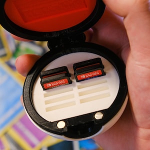 May include: A white and black round case with a red interior, open to reveal a white tray with slots for Nintendo Switch game cartridges. The case has a black rim and two silver magnets on the bottom.