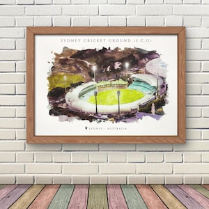 May include: A framed watercolor print of the Sydney Cricket Ground (SCG) in Sydney, Australia. The artwork depicts the stadium with a green field, stadium seating, and floodlights. The print is in a wooden frame and hangs on a white brick wall.