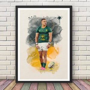 May include: A framed art print featuring a watercolour-style illustration of a rugby player in a green jersey and white shorts. The player stands with a rugby ball at their feet against a backdrop of grey and yellow ink splatters and text.