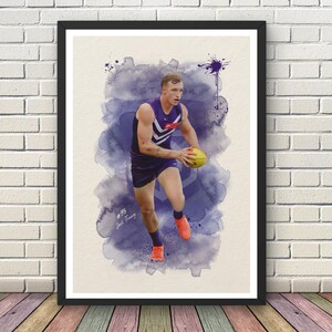 May include: A framed art print featuring a male athlete in a purple and white sleeveless jersey and shorts, holding a yellow football. The background is a purple watercolour splash effect with the text EST. 1994 and #35 Josh Treacy.