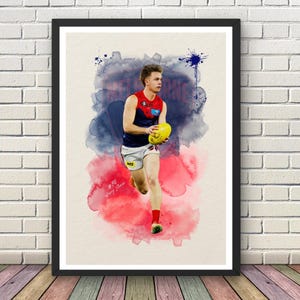 May include: A framed art print featuring a watercolor-style illustration of an Australian rules football player in a navy and red jersey and white shorts, holding a yellow football. The background has blue and red ink splashes and text.