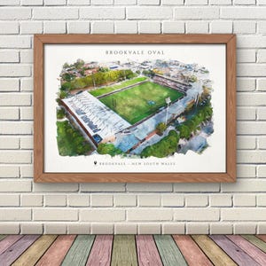 Könnte beinhalten: Ein gerahmter Aquarelldruck von Brookvale Oval in New South Wales. Das Kunstwerk zeigt eine Luftaufnahme des Sportplatzes mit grünem Rasen, Stadionbestuhlung und dem Text "BROOKVALE OVAL" oben.