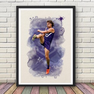 May include: A framed watercolour print depicting a football player in a purple kit kicking a ball. The background features a purple wash with the text "EST". The player wears orange shoes. The print is displayed on a wall.