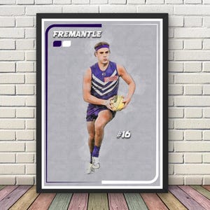 May include: Framed print with a watercolour effect depicting a football player in a purple and white uniform. The print includes the word "FREMANTLE" and the number "16". The player is holding a yellow football.