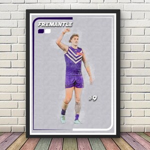 May include: Framed print with a watercolour effect of a Fremantle football player in purple and white kit. The player is celebrating with an arm raised. The print includes the team name and player number.
