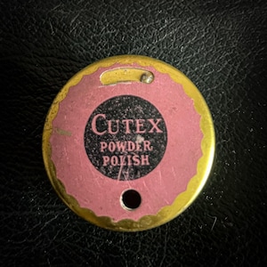 Könnte beinhalten: Ein runder, goldfarbener Metallbehälter mit einem rosafarbenen Deckel. Der Deckel hat einen schwarzen Kreis mit dem Text "CUTEX POWDER POLISH" in weißen Buchstaben.
