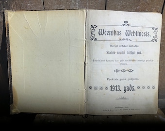 Op de afbeelding: Een oud, versleten boek met een titelpagina waarop "Weenibas Wehltnesis" staat. Het boek ligt open op de titelpagina en heeft een decoratieve rand rond de titel. De tekst op de titelpagina is in het Lets.