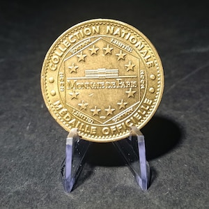 May include: A gold-colored coin with the inscription "Collection Nationale Medail Officielle" around the edge. The center of the coin features a building with the text "Monnaie de Paris" and eight stars.