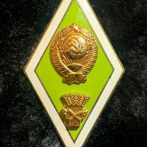 May include: A gold-colored metal pin with a green and white diamond-shaped background. The pin features a wreath with a hammer and sickle inside, and a sheaf of wheat below.