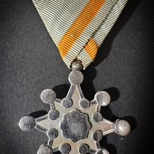 May include: A silver star-shaped medal with a textured center and a silver ribbon with two orange stripes.
