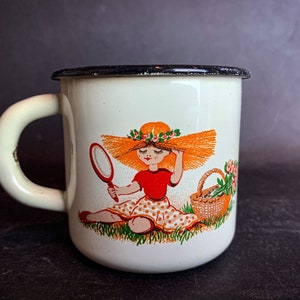 May include: White enamel mug with a black rim and a cartoon illustration of a girl wearing a straw hat and holding a mirror. The girl is sitting in a field with a basket of flowers.