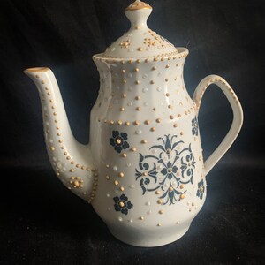 May include: White teapot with a gold and black floral design. The teapot has a lid and a handle. The design is made up of small dots and swirls.