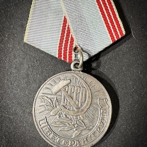 May include: A silver medal with a red star and hammer and sickle, surrounded by a laurel wreath. The medal has the text "СССР" and "ВЕТЕРАН ТРУДА" inscribed on it. The medal is attached to a gray and red ribbon.