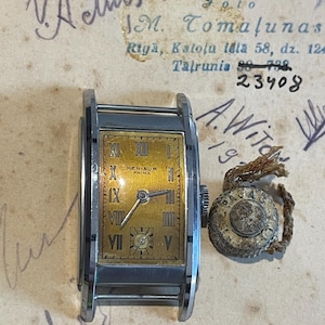 May include: Vintage rectangular wristwatch with a gold-tone face and Roman numeral hour markers. The watch, marked "KENISUR PRIMA," has a silver-tone case and is accompanied by a separate winding crown. The background features handwritten notes and the number 23408.