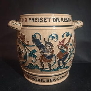 May include: A ceramic beer stein with two handles, featuring a scene of two men carrying large barrels. The stein is decorated with floral designs and the text "Preiset die Reben Wohl Bekomm".
