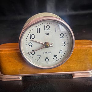 May include: A white analog clock with black numbers and hands, set on a wooden base. The clock face reads "USSR" and "Bocxoo".