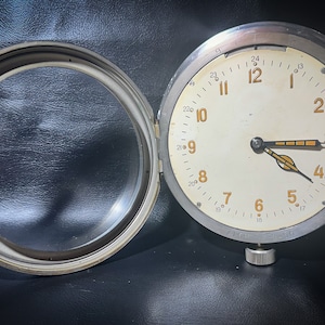 Könnte beinhalten: Eine silberne Uhr mit weißem Zifferblatt und schwarzen Zeigern. Die Uhr hat eine runde Lünette und einen kleinen Knopf an der Unterseite. Die Uhr ist geöffnet und das Glas des Zifferblatts ist vom Uhrwerk getrennt.